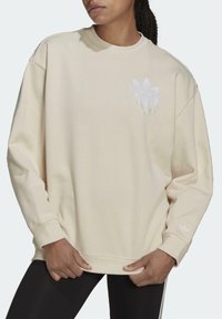 Beige sweatshirt made of soft fabric, featuring a white embroidered logo with fringe detail, long sleeves, and a classic crew neck design.