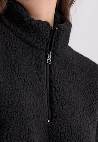 Svart fleece halv-zip genser med høy krage, strukturert overflate og detalj med sølv glidelås. Mykt, varmt materiale.