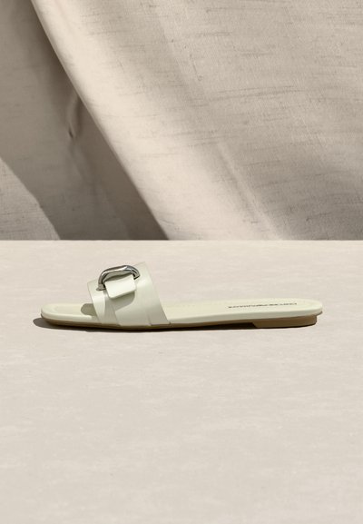 Płaski biały sandał wsuwany z szerokim paskiem i srebrną klamrą, wyeksponowany na jasnobeżowej powierzchni z tkaninowym tłem.