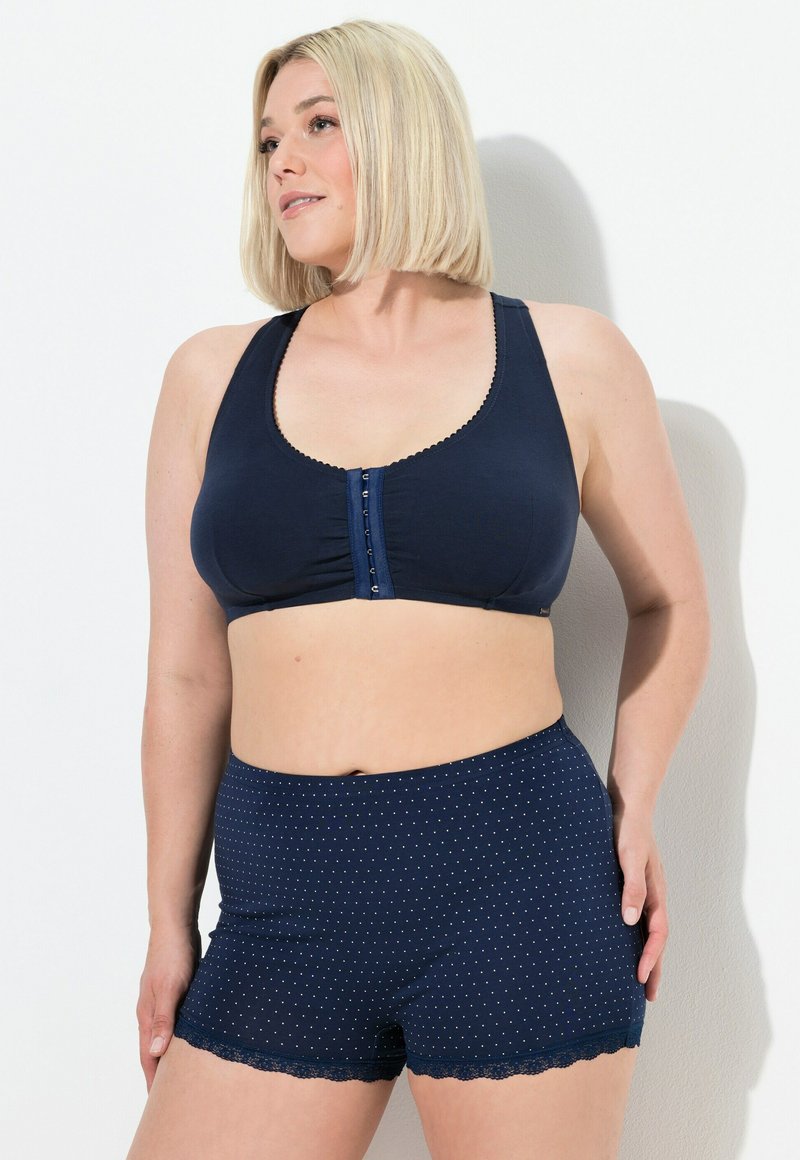 Donna con capelli biondi corti, che indossa un reggiseno blu scuro con chiusura frontale e pantaloni corti blu scuro con pois e bordo in pizzo, in piedi di fronte a un muro bianco.