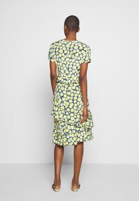 Una modella indossa un vestito floreale con motivi gialli e blu, con rouches all'orlo, e sta in piedi con la schiena rivolta verso l'osservatore.