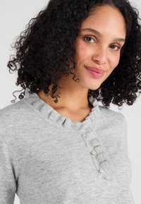 Maglione grigio chiaro con scollatura arricciata, caratterizzato da tre bottoni decorativi e una texture morbida. Primo piano dell'area del colletto.