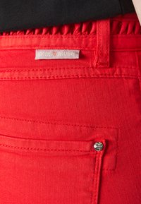 Jeans de ganga vermelhos com um botão prateado e um remendo de etiqueta. O tecido tem uma textura suave com detalhes de costura visíveis e uma cintura ajustada.