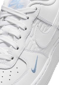 Valkoiset Nike-tennarit, joissa on sileä nahka, tekstuuriset yksityiskohdat ja vaaleansinisellä brodeerattu logo. Nauhat ovat valkoiset ja niissä on tavallinen reikämalli.