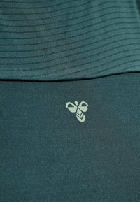 Teal textil med ribbat mönster; inkluderar en liten ljusgrön bi-logotyp längst ner, vilket framhäver brandingdetaljen och designen.