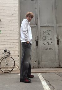 En person står i hettegenser i hvitt, løse grå jeans og svart-røde sko. En sykkel lener mot en vegg, og graffiti er synlig på døren.