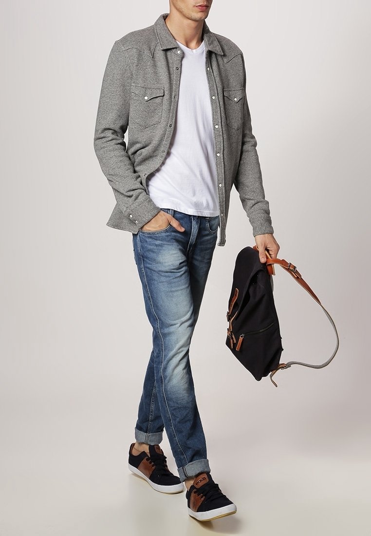 Homme portant une veste-chemise grise boutonnée, un t-shirt blanc, un jean bleu, des baskets noires, tenant un sac à dos noir par la lanière.