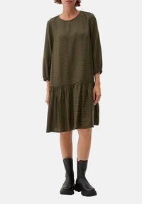 Langärmliges, olivgrün kariertes Kleid mit gestuften Rüschensaum, aus weichem Material gefertigt. Kombiniert mit schwarzen, klobigen Stiefeln.