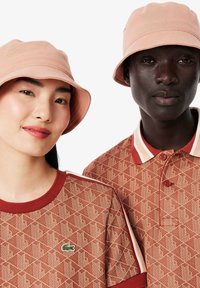 Dos personas llevan gorras de cubo de color rosa claro a juego. La camisa de la mujer es de textura, marrón con un patrón geométrico y un pequeño logo. La polo del hombre tiene cuello y un patrón marrón similar.