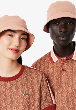 Zwei Personen tragen passende hellrosa Bucket Hats. Das Shirt der Frau ist strukturiert, braun mit einem geometrischen Muster und einem kleinen Logo. Das Polo des Mannes hat einen Kragen und ein ähnliches braunes Muster.