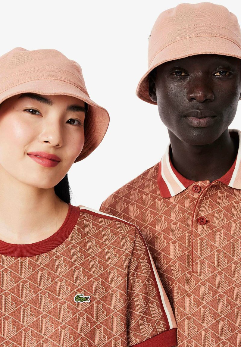 Dos personas llevan gorras de cubo de color rosa claro a juego. La camisa de la mujer es de textura, marrón con un patrón geométrico y un pequeño logo. La polo del hombre tiene cuello y un patrón marrón similar.