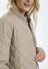 Veste bomber matelassée beige avec un col côtelé, fermeture éclair et poches latérales. Présente un motif en vagues subtil et une texture lisse et brillante.