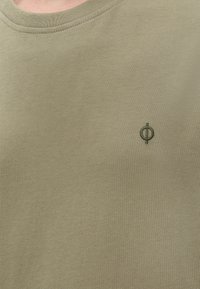 Gros plan d’un t-shirt vert kaki à col rond avec un petit logo circulaire vert foncé brodé sur le côté gauche de la poitrine.