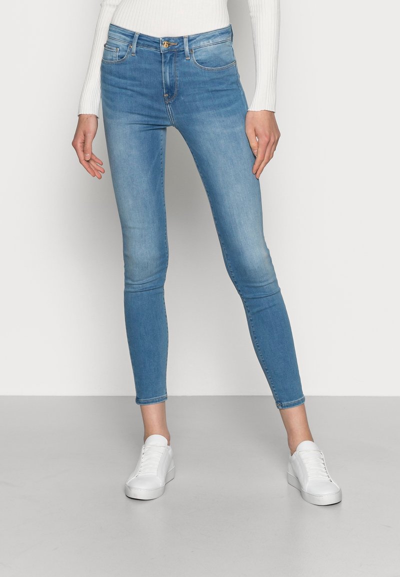 Mujer vestida con jeans skinny azul claro, top de manga larga blanca con canalé y zapatillas blancas, de pie contra un fondo claro y uniforme.
