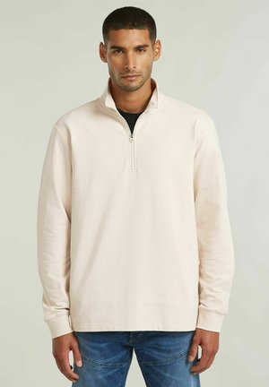 Man draagt een lichtbeige pullover met kwart-ritssluiting, een zwart shirt eronder en blauwe spijkerbroek, staand tegen een effen lichte achtergrond.