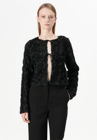 Veste noire courte avec des appliqués floraux texturés, devant ouvert avec un lien de fermeture et manches longues, associée à un pantalon noir.