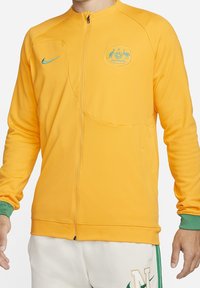 Nike zip-jakke i lys gul, med en grøn detalje på manchetterne, et logo-patch med kænguruer og en glat, strækbar stoftekstur.