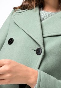 Žena v svetle zeleném dvojitém vlněném kabátu s černými knoflíky přes šedý pletený svetr, upravuje rukáv kabátu.