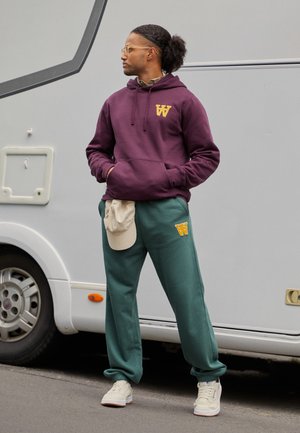 Burgunderfarget hettegenser med gul logo, grønne joggebukser, beige caps hvilende på livet, og hvite sneakers. Naturlig hår stylet bakover.
