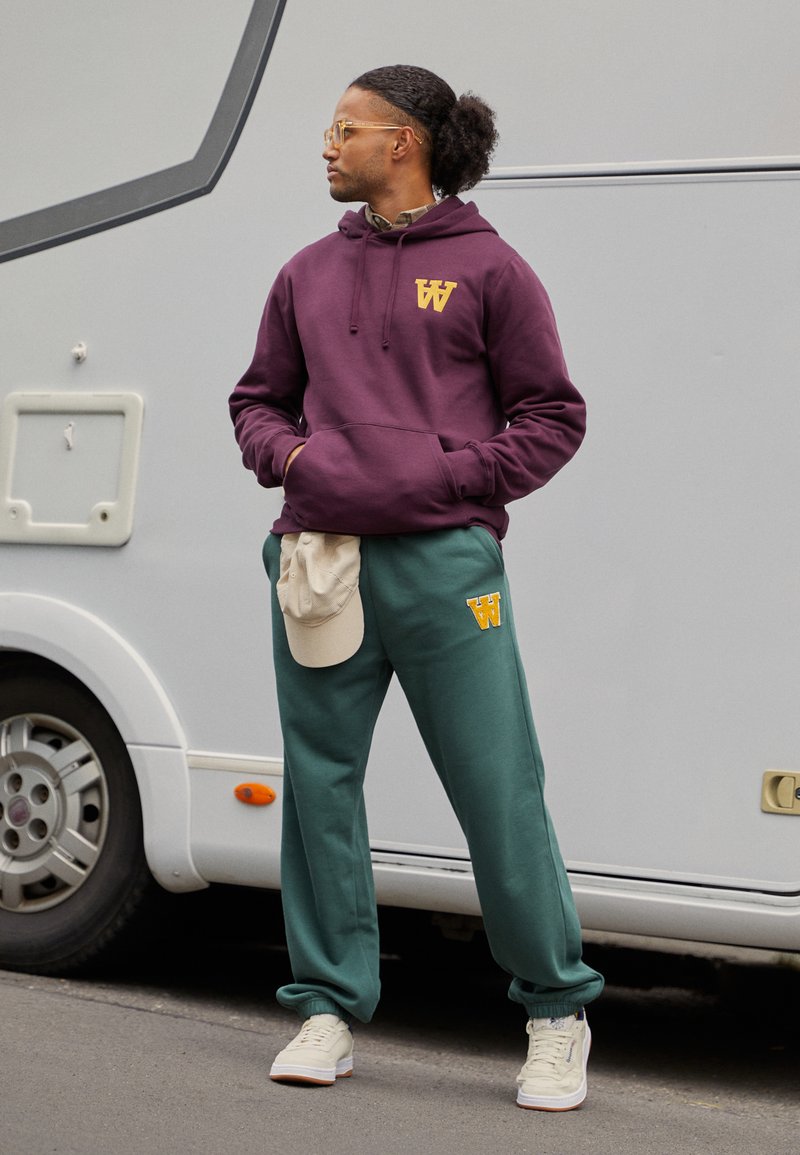 Vinröd huvtröja med gul logotyp, gröna träningbyxor, beige keps vilande på midjan och vita sneakers. Naturligt hår stylat bakåt.