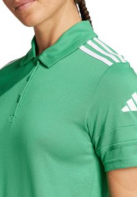 Polo shirt verde con un tessuto testurizzato, colletto pieghevole, tre strisce bianche sulla spalla e un dettaglio logo sottile.