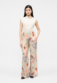 Γυναίκα στέκεται φορώντας αμάνικη μπεζ μπλούζα και παντελόνι με φαρδιά μπατζάκια με πολύχρωμα λουλουδένια σχέδια και καφέ παπούτσια, με τα χέρια στις τσέπες.