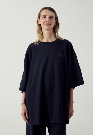 Ung kvinde med blondt hår iført en oversize sort t-shirt og matchende bukser, stående foran en ensfarvet lys baggrund.