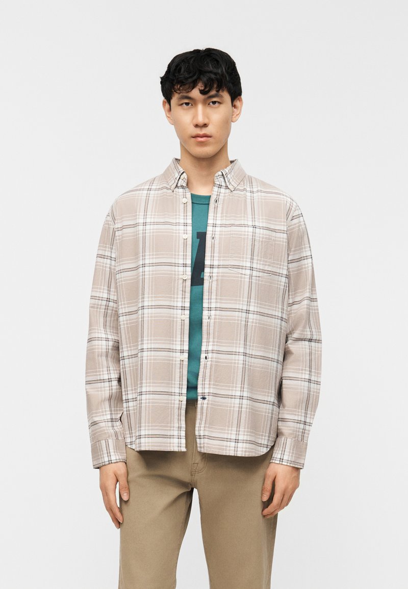 Νεαρός άνδρας που φοράει μπεζ πουκάμισο με καρό κουμπωμένο πάνω από πράσινο μπλουζάκι με γραφικό σχέδιο και χακί παντελόνι, στέκεται μπροστά σε απλό λευκό φόντο.