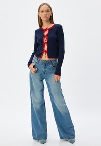 Cardigan a coste blu navy con rifinitura rossa, caratterizzato da un dettaglio con nodo sulla parte frontale. Abbinato a jeans a gamba larga azzurri chiari e tacchi a punta color teal.