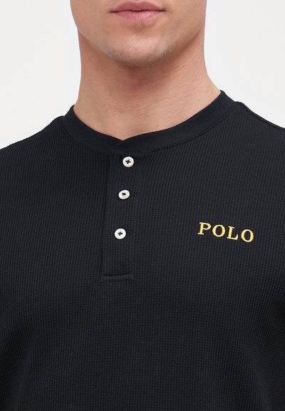 Polo shirt preto em malha waffle com três botões brancos, apresentando bordado dourado de "POLO" no peito esquerdo. Design de colarinho redondo.