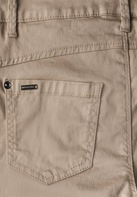 Béžové látkové kalhoty s malou zadní kapsou opatřenou kovovou logovou štítkem, čistým šitím a hladkou texturou.