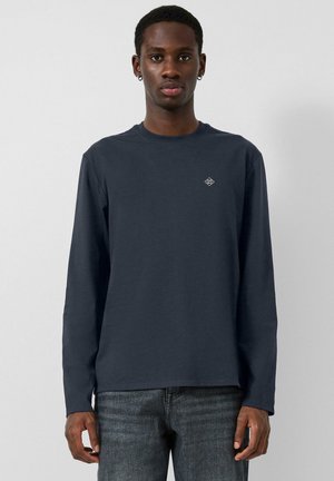 Jonge man die een donker shirt met lange mouwen draagt met een klein logo op de borst en een donkere spijkerbroek, gericht naar voren tegen een effen achtergrond.