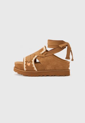 Sandalias de ante marrón con tiras cruzadas, un patrón decorativo, lazos en el tobillo y suela plataforma texturizada. Presenta un diseño resistente y plano.