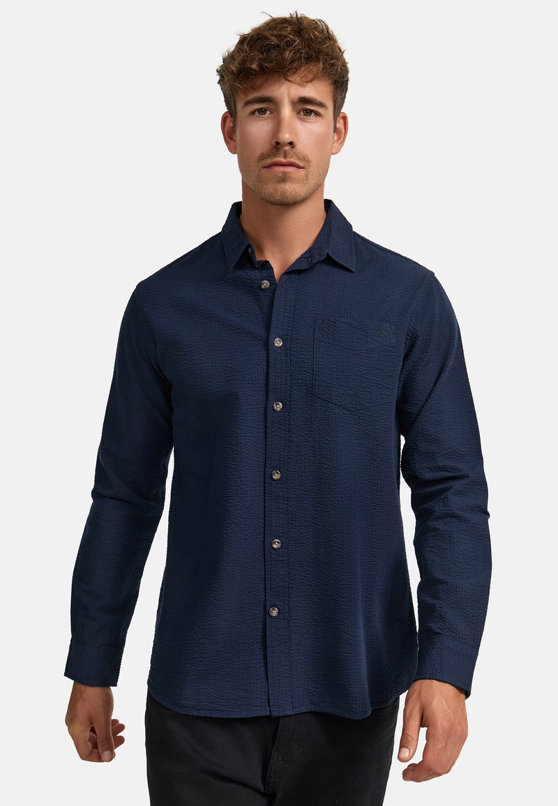 Homme aux cheveux courts et bouclés portant une chemise bleu foncé texturée à manches longues avec une poche poitrine, debout devant un fond uni.
