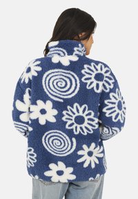 Una donna vista da dietro che indossa una giacca in pile blu con motivi a spirale bianchi e fiori, abbinata a jeans azzurri chiari.
