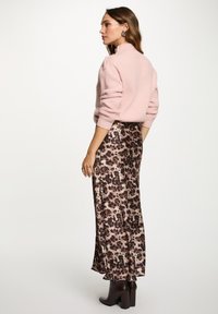 Vrouw in een lichtroze trui, satijnen rok met luipaardprint en donkerbruine enkellaarsjes, staand in profiel tegen een effen achtergrond.