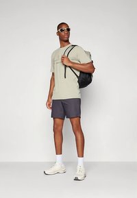 Muškarac u naočalama za sunce, bež majici, tamnim kratkim hlačama, bijelim čarapama, bijelim tenisicama i nosi crni ranac preko jednog ramena.