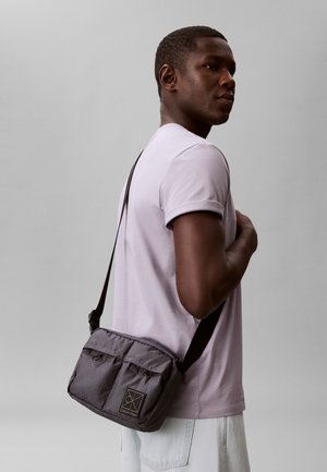 Joven con camiseta lila claro que lleva un bolso bandolera gris de Calvin Klein sobre el hombro, con un fondo liso.