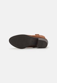 Chaussure en daim marron avec une tige à motif, dotée d'une semelle en caoutchouc noir texturé et d'un petit talon. Accent de boucle près de la cheville.