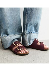 Burgundy mockasksandaler med dubbla guldfärgade spännen, korksula och en öppen design. Bärs med lösa, uppvikta blå denimjeans.