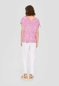 Rosa, blumengemusterte Bluse mit kurzen Ärmeln, V-Ausschnitt und Bindebändern an den Ärmeln, kombiniert mit weißen, schmal zulaufenden Hosen und beigem Schuhwerk.