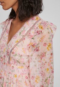 Blomstrete kjole i rosa stoff, med volanger på ermene, delikat blonder, og flerfargede blomsterdesign med grønne detaljer.
