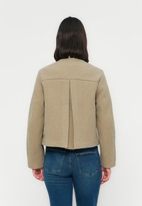 Kvinne med rett mørkt hår iført en beige kort jakke og blå jeans, sett fra ryggen mot en ensfarget hvit bakgrunn.