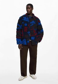 Farverig fleecetrøje med abstrakte mønstre i blå, rød og sort, med knaplukning, stor pasform og høj krave. Kombineret med brune bukser.