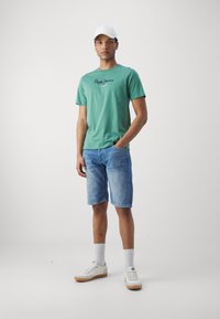 Zöld pamut póló logóval, farmer rövidnadrág, fehér sportcipő és fehér sapka. A modell az egyik kezét a zsebében tartja, semleges háttér előtt.