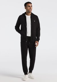 Zwarte ritshoodie met kangoeroe pockets, gecombineerd met zwarte joggerbroek. Materiaal lijkt zacht; hoodie heeft een logo op de borst. Witte sneakers.