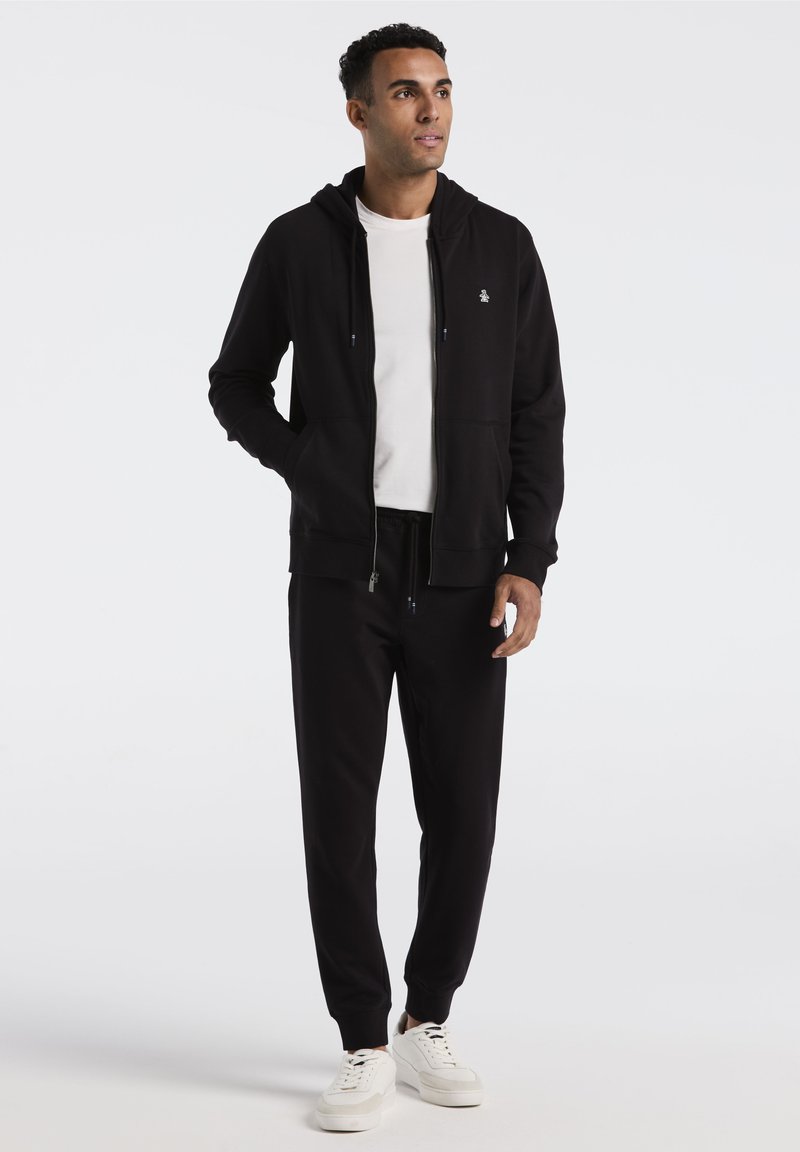 Zwarte ritshoodie met kangoeroe pockets, gecombineerd met zwarte joggerbroek. Materiaal lijkt zacht; hoodie heeft een logo op de borst. Witte sneakers.