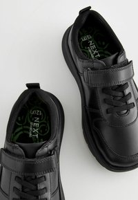 Zwarte synthetische schoenen met een gladde afwerking, voorzien van een klittenbandsluiting en traditionele veters, en groene binnenzolen met een logo-patroon.