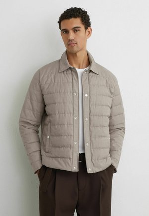Junger Mann trägt eine hellbeige gesteppte Jacke über einem weißen Hemd und dunkelbraunen Hose, steht mit den Händen in den Taschen vor einer schlichten Wand.