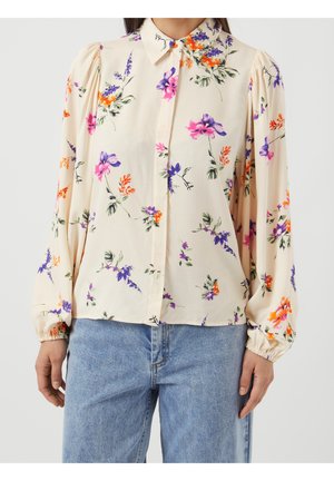 Femme portant un chemisier crème à manches longues avec imprimé floral coloré et poignets boutonnés, associé à un jean en denim bleu clair.